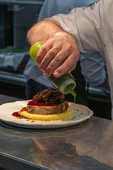 Finition au spray d'huile — Coulisses du Bistrot 270. Chef en veste blanche pulvérisant un spray d'huile verte sur un plat au pass du Bistrot 270
